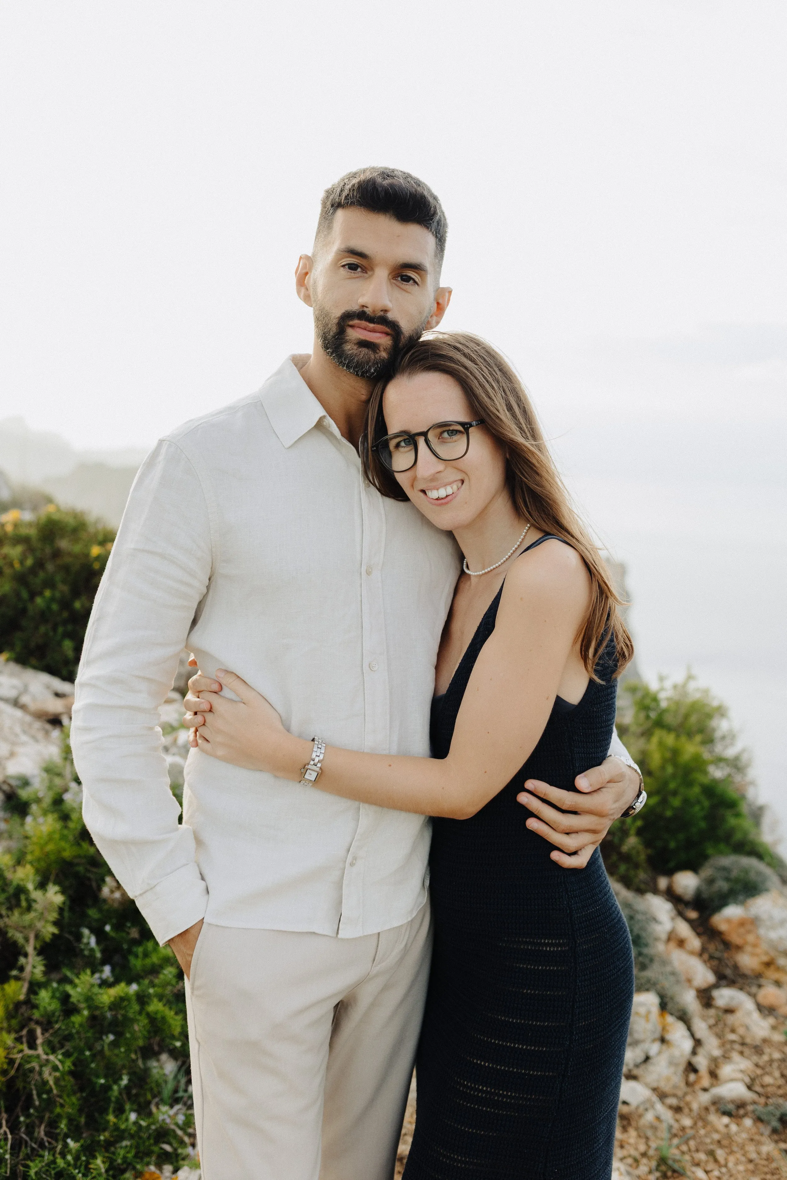 A couple in a cream shirt and a dark dress leaning into each other above the coast