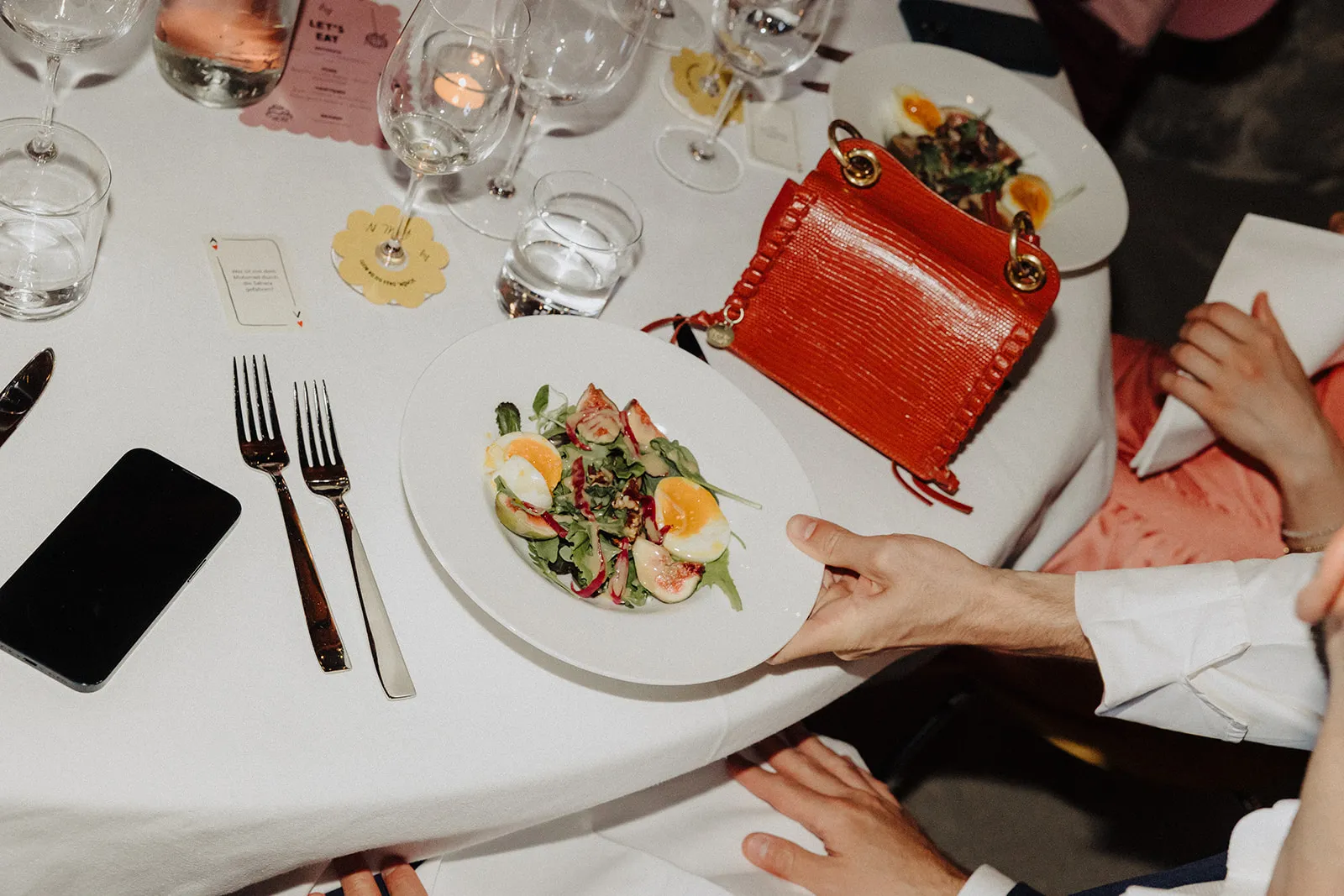 A plate of salad and a red handbag on a laid table, hands reaching in