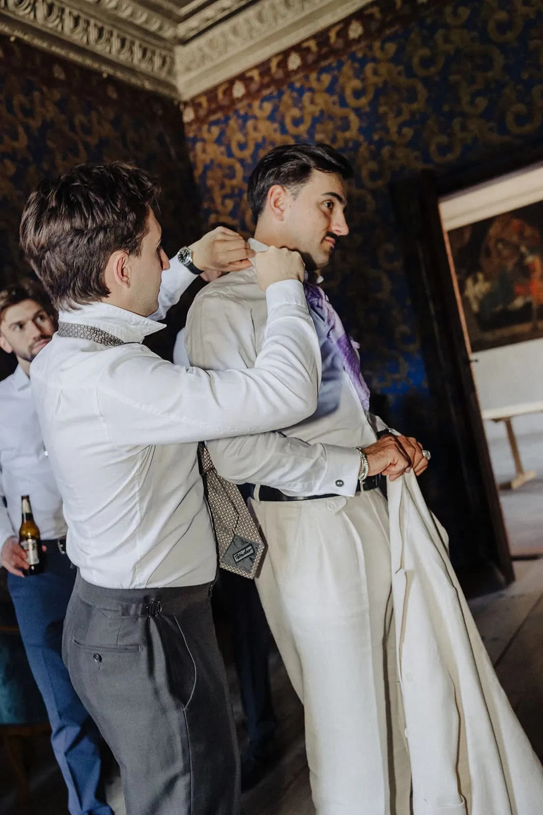 A friend doing up the groom's collar while he holds his trousers