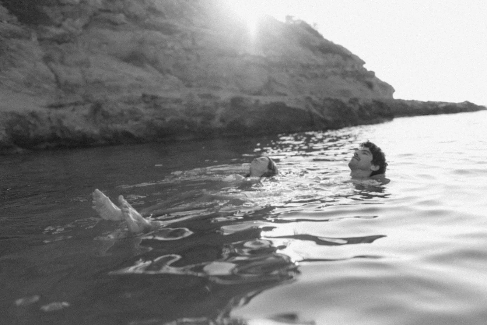 Two people swimming in the sea below a headland, the sun low