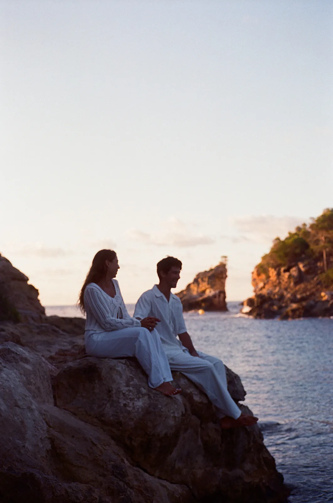 A couple sitting on the rocks above the water at dusk