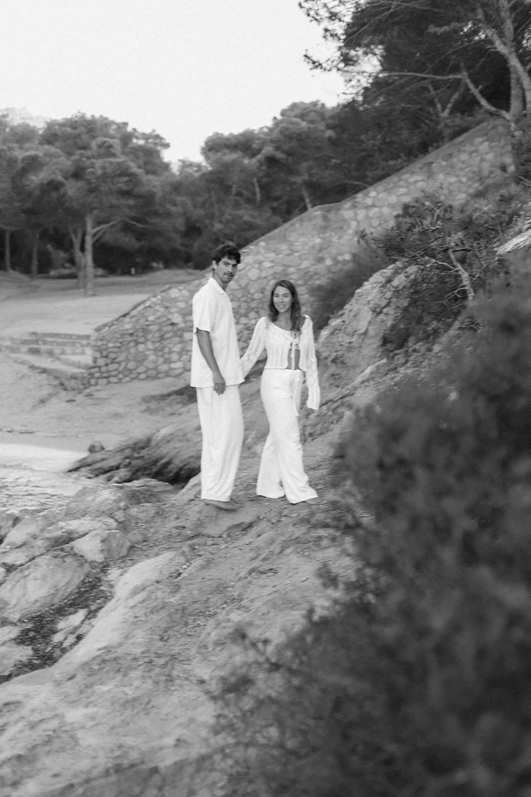 The couple in white walking hand in hand over the rocks, pines behind them