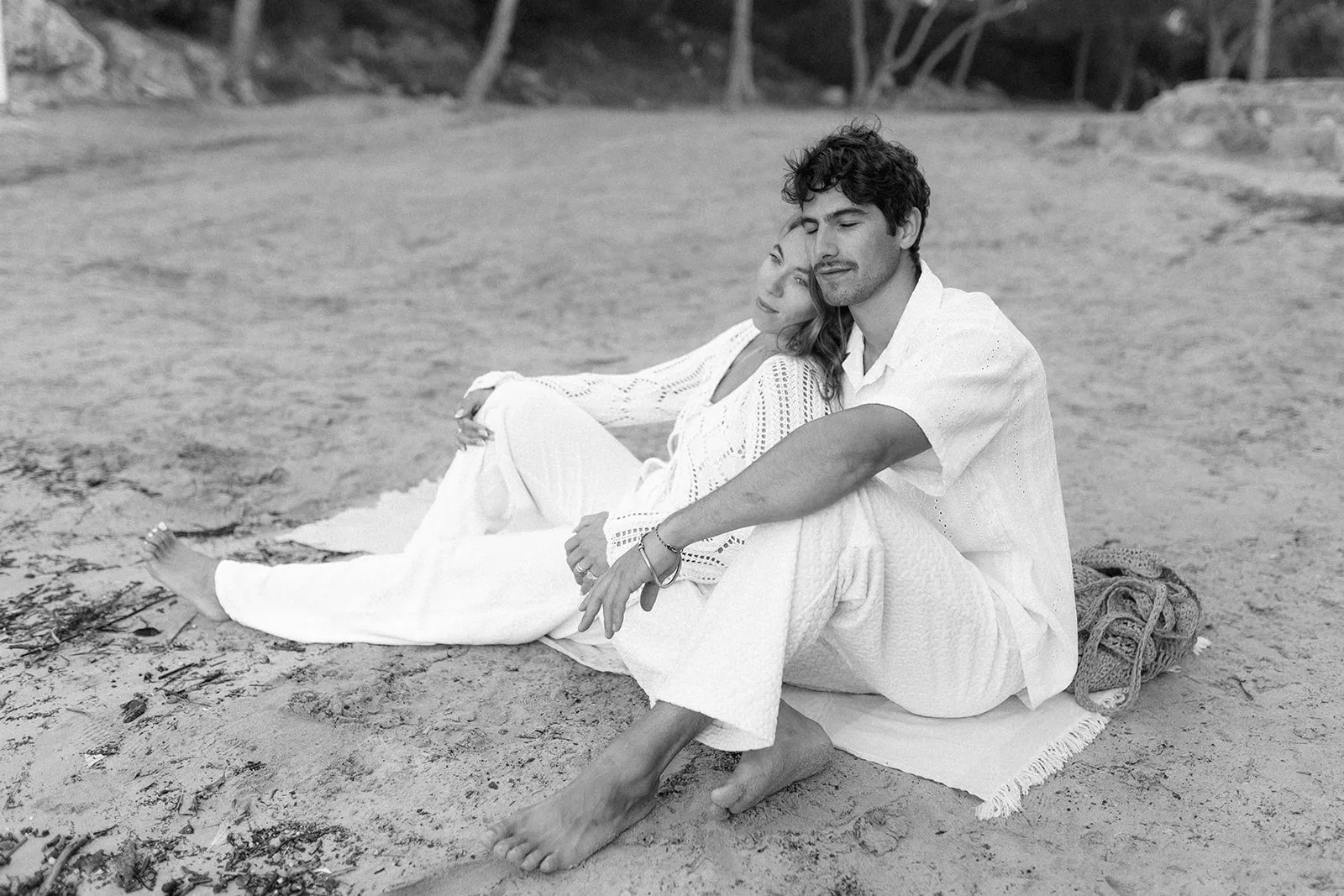 A couple sitting on the sand in white, leaning together