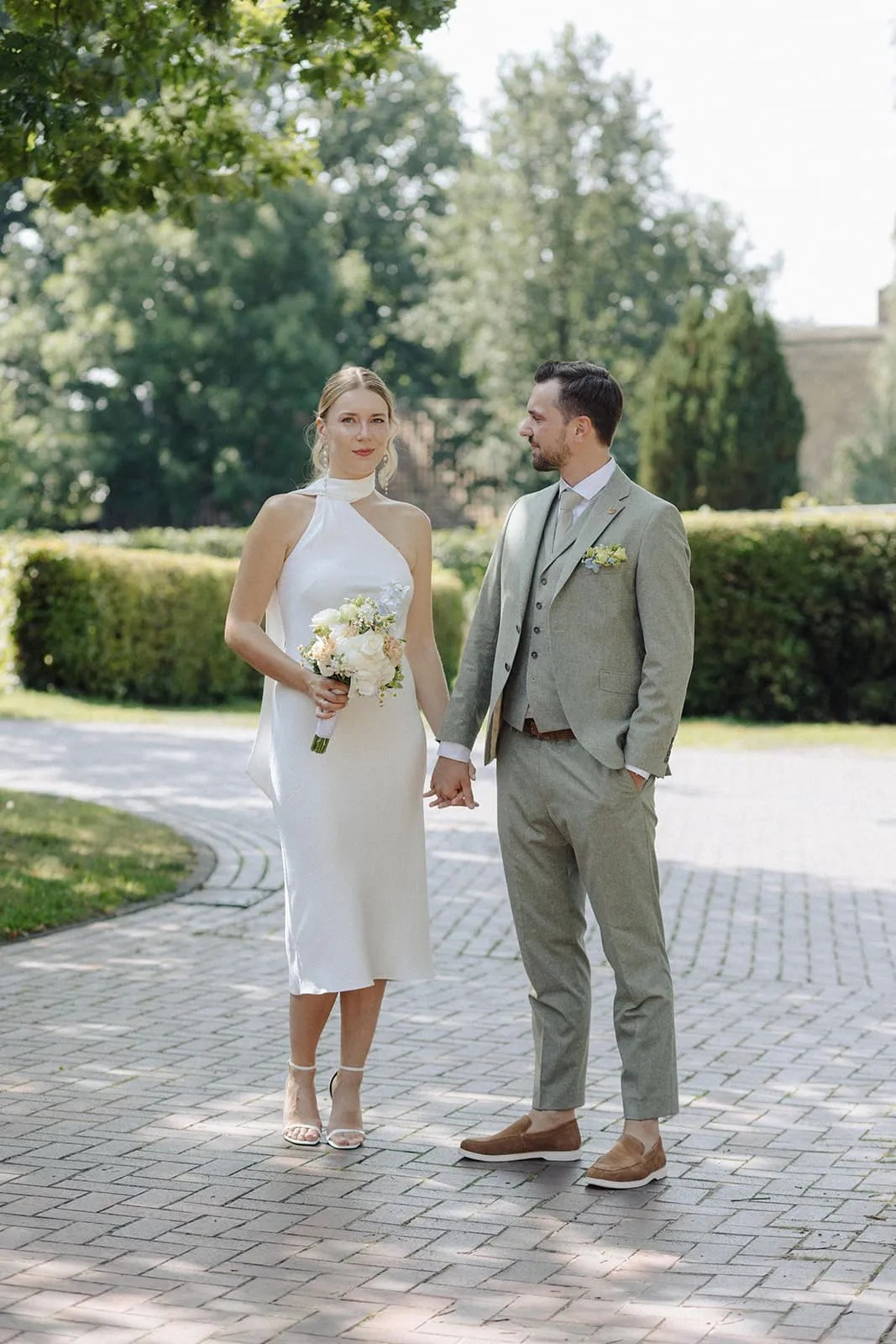 The couple standing on a park path, looking at each other, clipped hedges behind