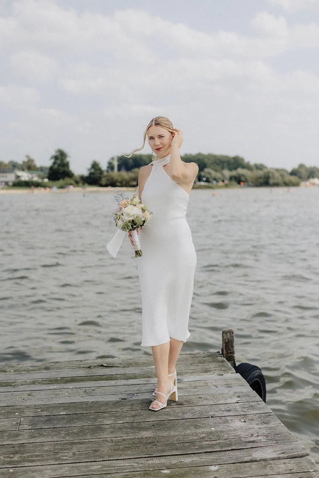 The bride alone on a wooden jetty, bouquet in hand, the lake behind her