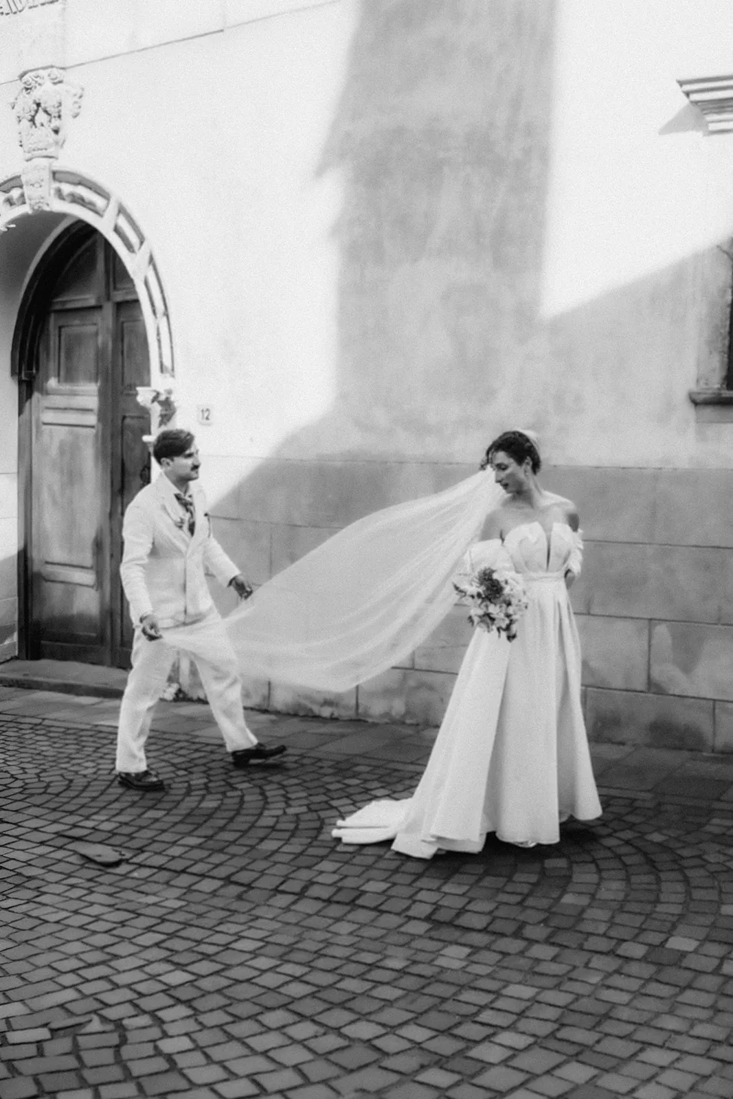 The couple walking across cobblestones, the veil carried behind them