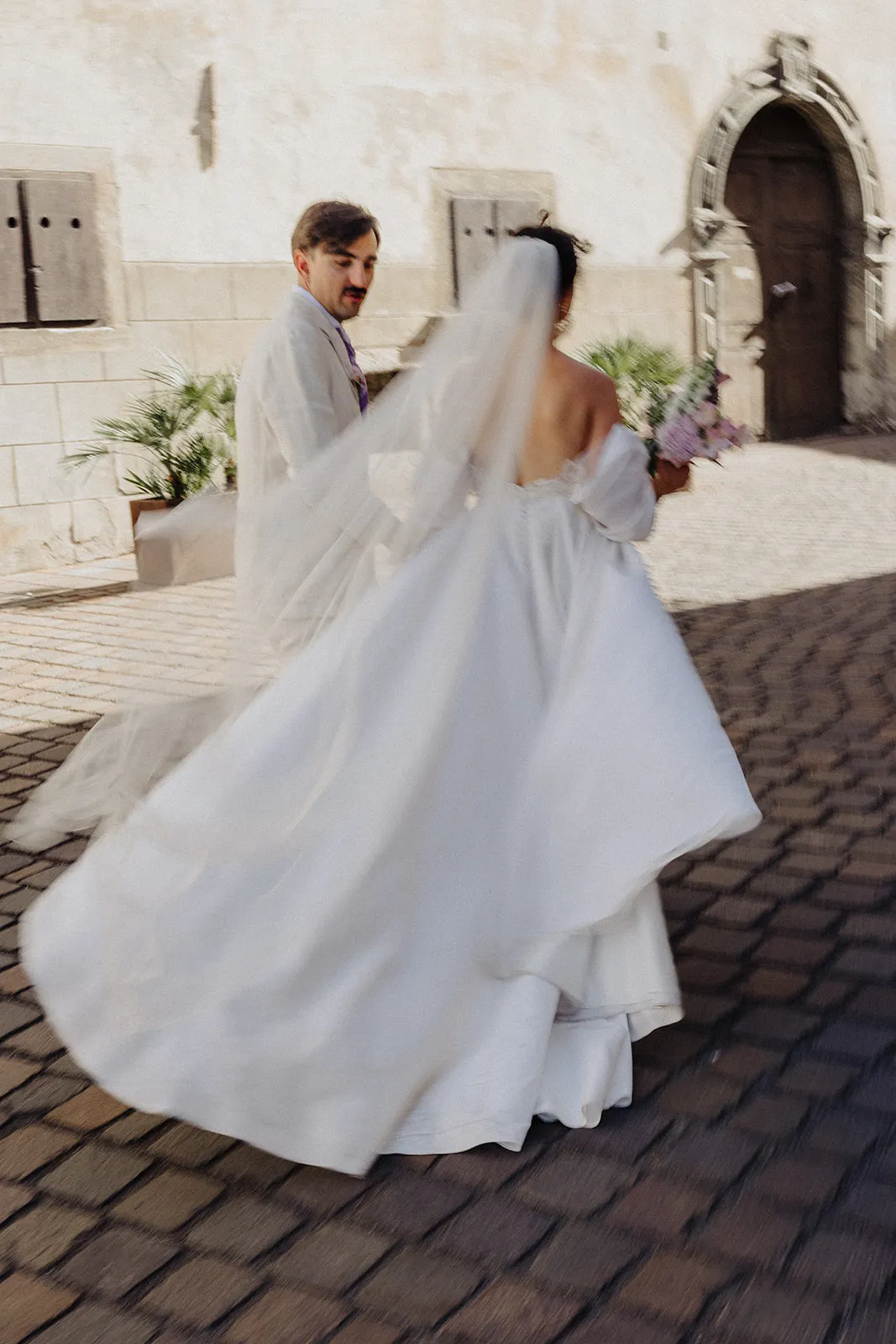 Die Braut dreht sich, Kleid und Schleier schwingen mit, der Bräutigam schaut ihr auf dem Pflaster in Margreid zu.