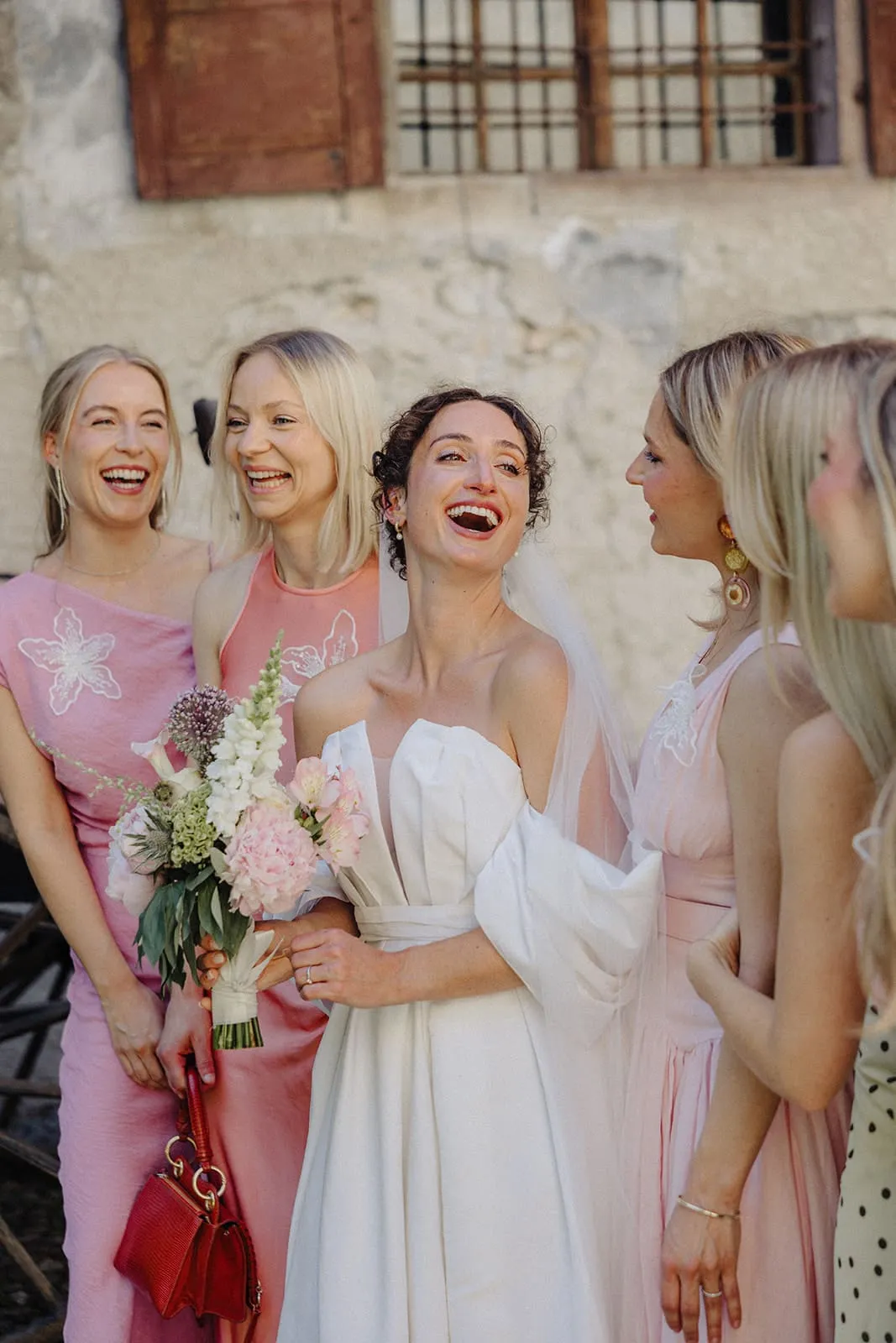 The bride laughing among her friends in pink in front of the stone wall